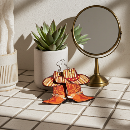 Cowboy boot earrings on a tiled surface with a plant and mirror in the background.