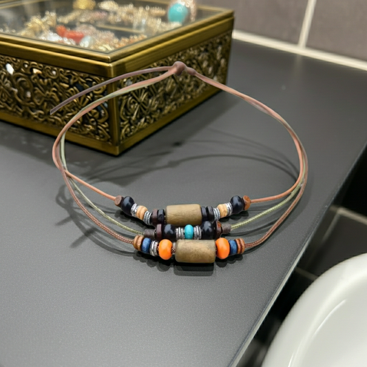 A set of three thin cord necklaces with central cylindrical beads and multi-colored spacer beads, displayed on a dark gray surface with an ornate gold jewelry box in the background.