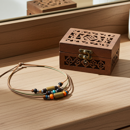 A small carved wooden jewelry box with a brass clasp sits on a light wood surface beside a beaded cord necklace with colorful spacer beads.
