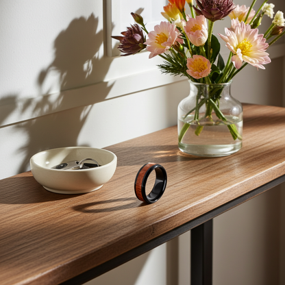A black magnetic ring with a reddish-brown wood inlay stands on a wooden console table next to a small cream ceramic bowl with keys and a glass vase of flowers.
