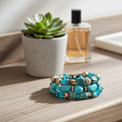 A stack of three beaded bracelets with turquoise, blue, and gold beads, styled on a light wood surface next to a small potted succulent and a perfume bottle.
