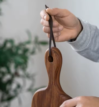 Wooden cutting board with handle held by a person against a blurred green plant background