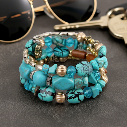 A stack of three beaded bracelets featuring turquoise-colored stones, metallic gold and silver beads, and a brown wooden bead, displayed on a dark textured surface with sunglasses and keys in the background.