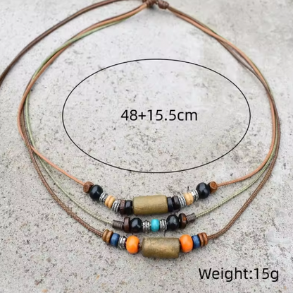 A multi-strand cord necklace with decorative beads and two central metallic cylindrical charms, shown on a light gray textured surface. Black oval line art and text indicate a size of 48+15.