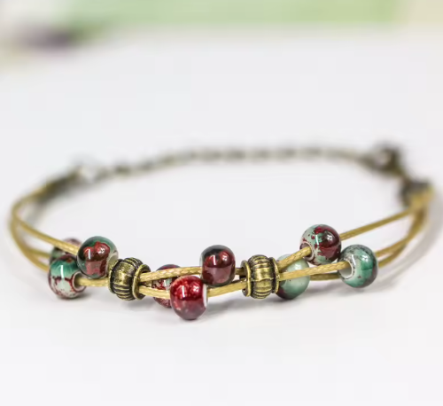 A close-up of a delicate bracelet with multiple strands of antique gold-toned wire and glossy glass beads in deep red, green, and marbled tones, set against a soft, out-of-focus white background.