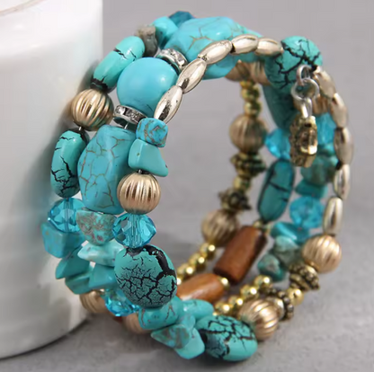 A close-up of a multi-strand beaded bracelet featuring turquoise blue stones with black matrix cracks, metallic gold and silver beads, and a small wooden bead accent, shown next to a white ceramic object on a gray surface.